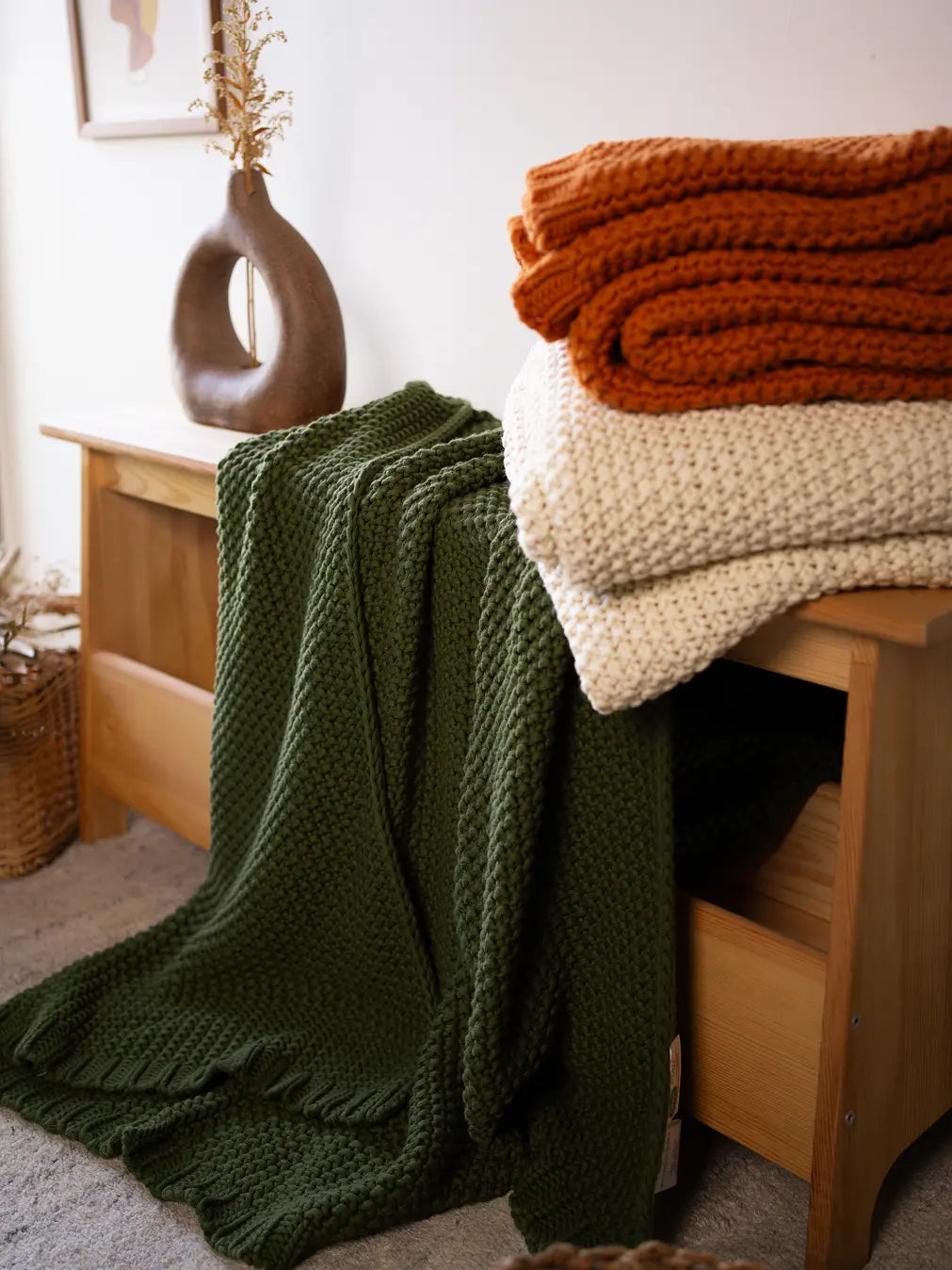 Alpine Cotton Knitted Throw in green, off-white, and mustard, neatly stacked on a wooden bench.