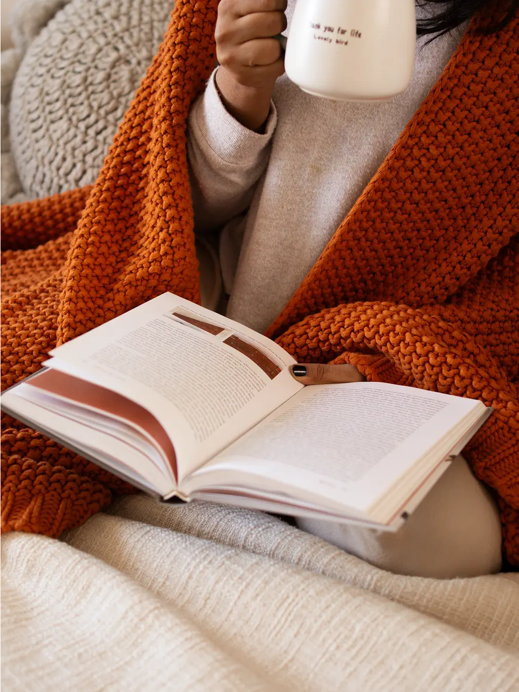 Person wrapped in Alpine Cotton Knitted Throw, enjoying a book and a warm drink in a cozy setting