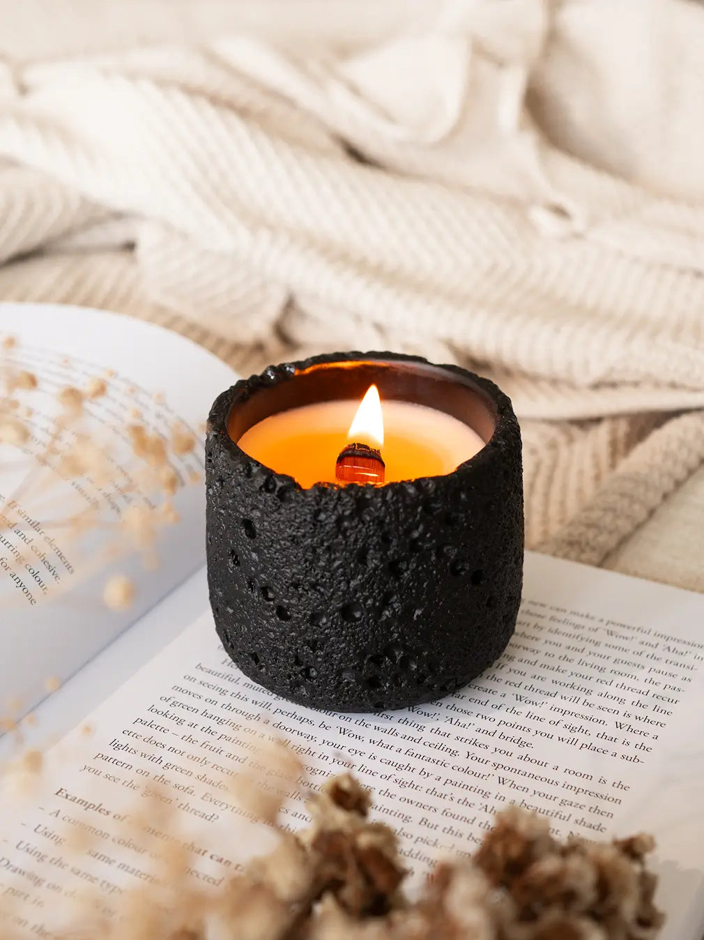 Capri Concrete Scented Candle in black burning on an open book with a soft blanket in the background