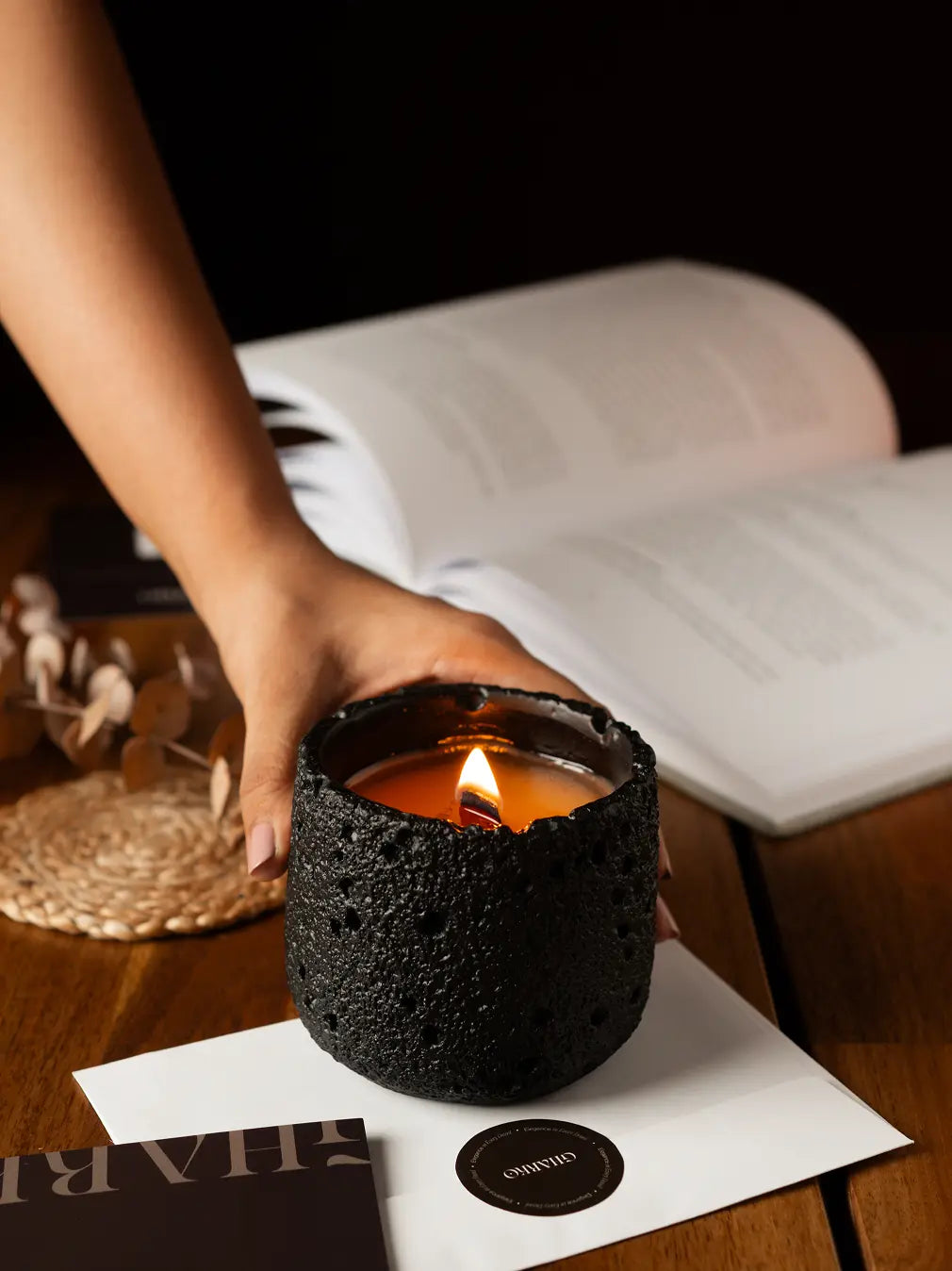 Hand holding a lit scented candle on a wooden surface with a book in the background.