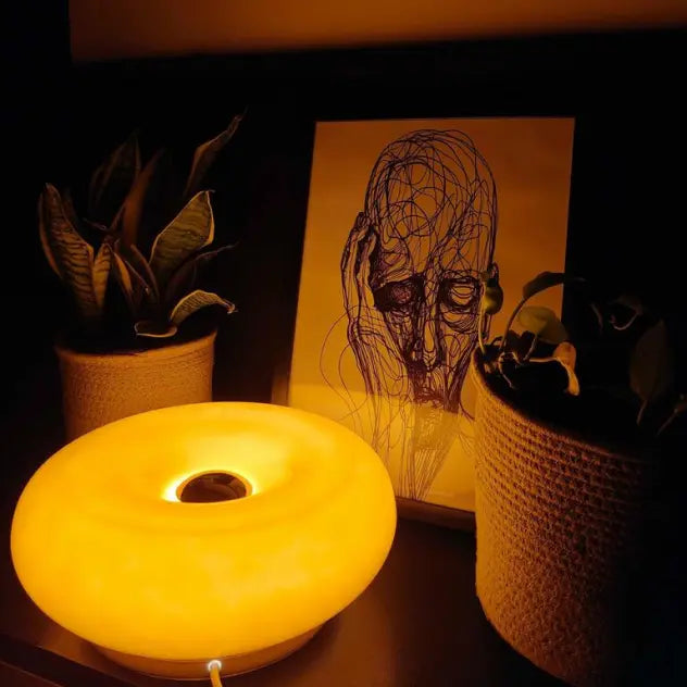 Round Orange Lamp on a dark surface with plants and a framed drawing in the background.