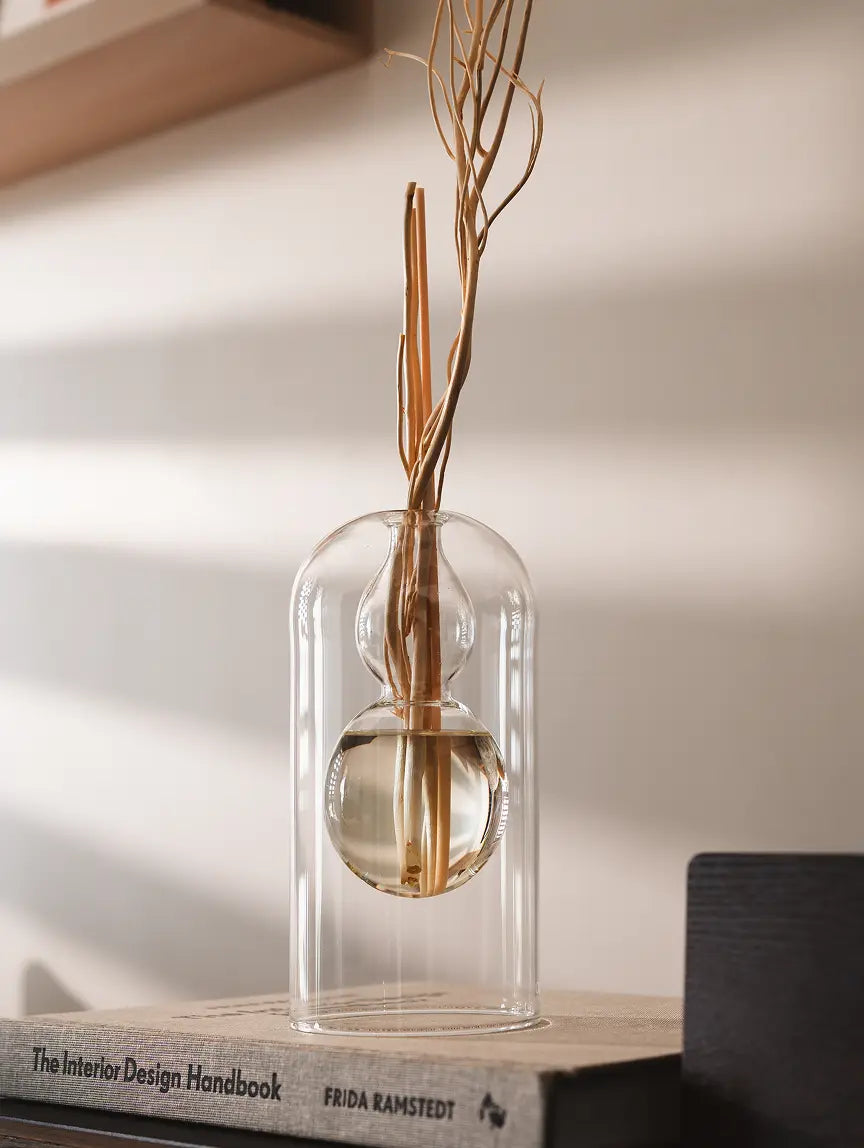 Transparent Calmma Glass Reed Diffuser with rattan reeds, placed on design books, reflecting natural light.