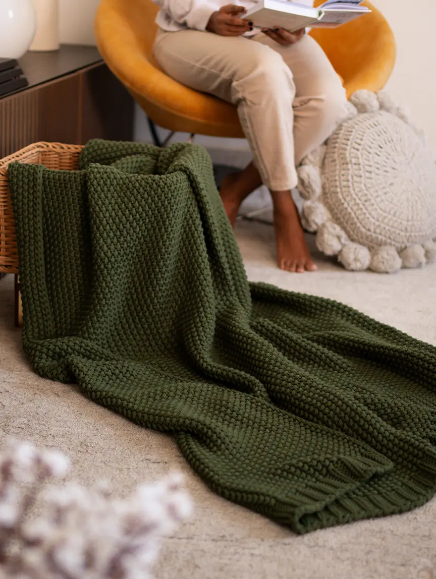 Green Alpine Cotton Knitted Throw draped over a basket, adding warmth to a cozy reading nook