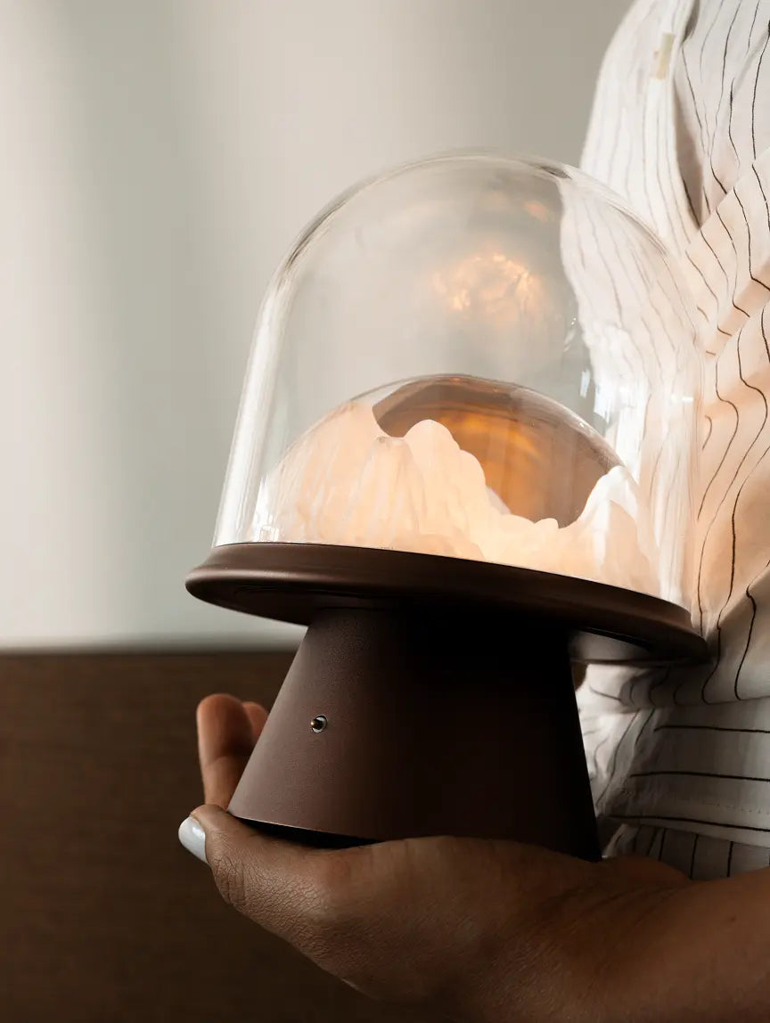 Hand holding a small wireless lamp with a glass dome and brown metal base.