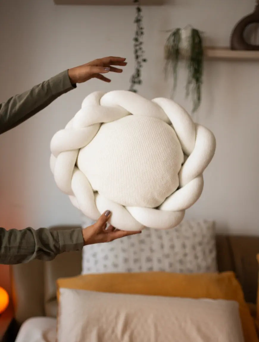 Gharko White sculptural cushion with braided edge being held above cozy bed setup