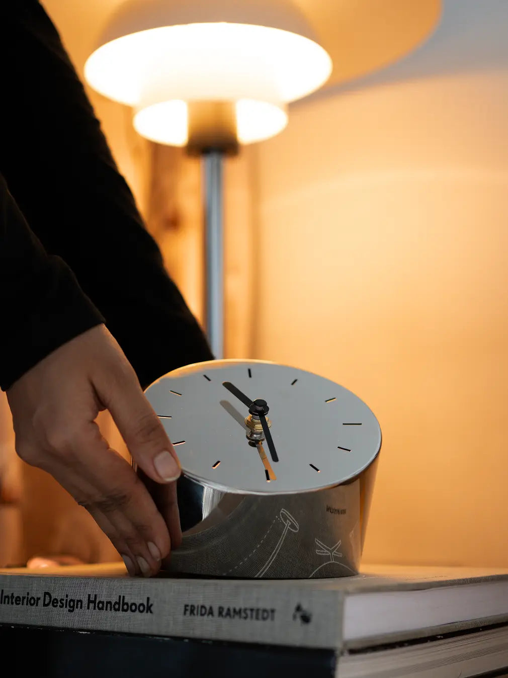 Polished metal table clock being placed near lamp on bedside stacked with books