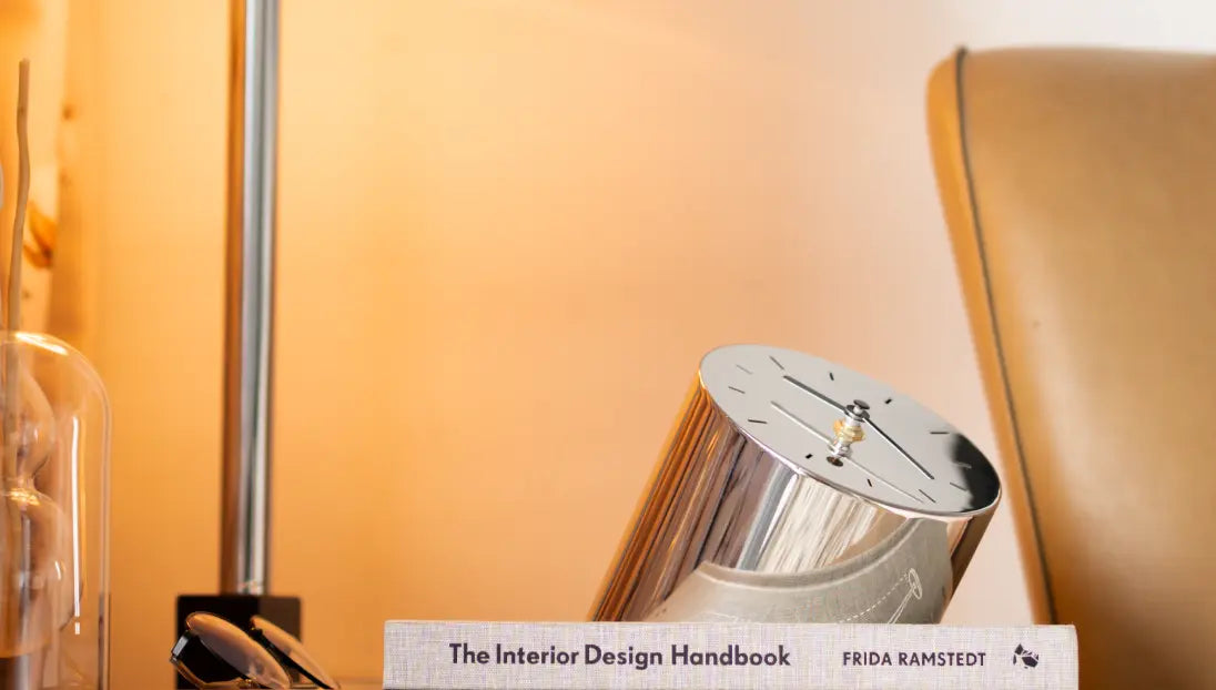 Sculptural metal table clock with mirror finish leaning against books under soft lighting