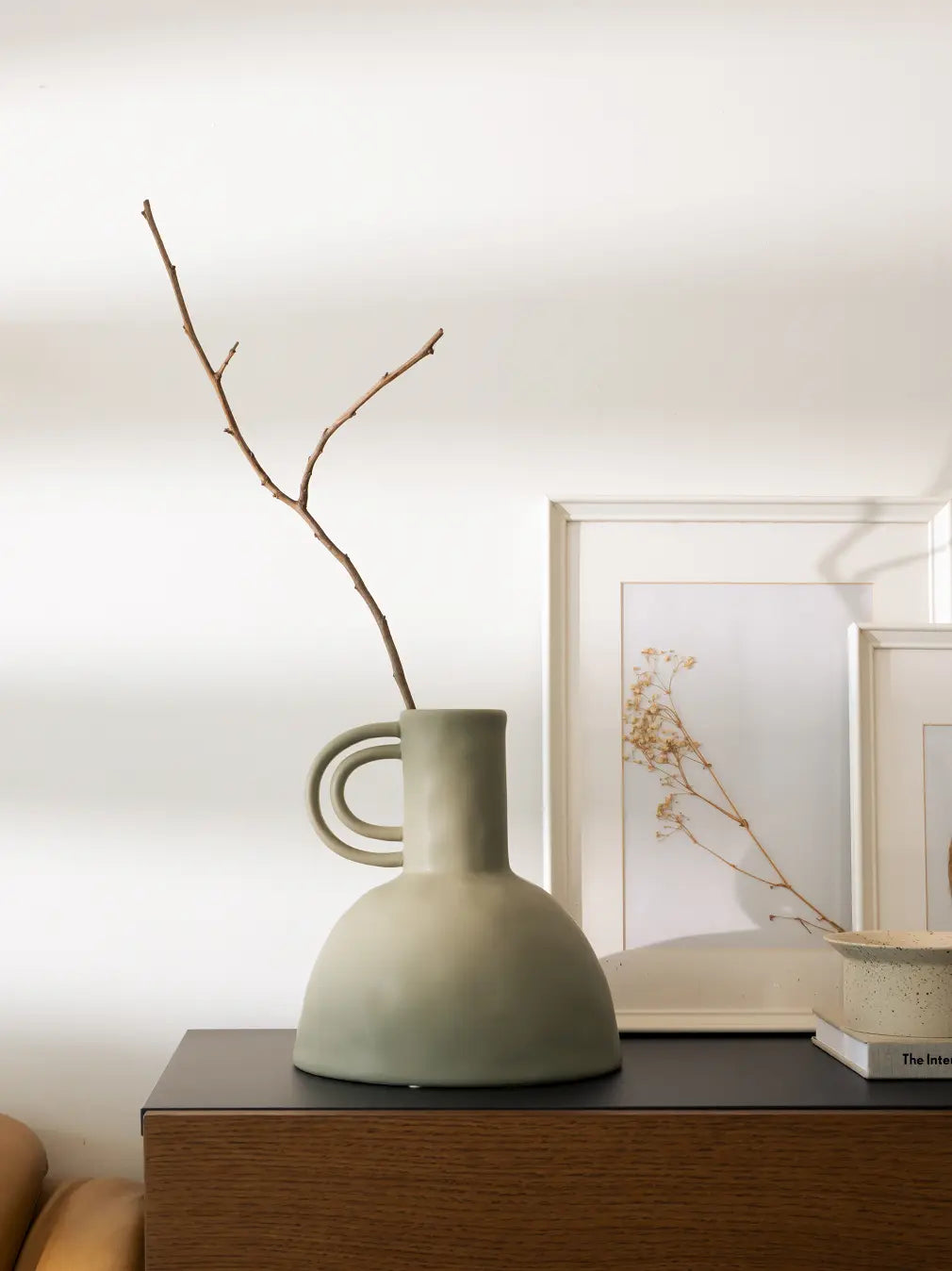 Green ceramic vase with a branch on a wooden surface, framed artwork in the background
