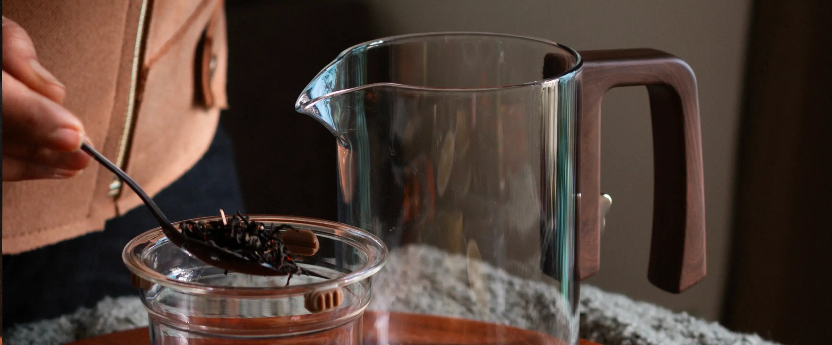 Pourfect Tea Infuser with a clear glass body, wide spout, and ergonomic wooden handle, as loose tea leaves are added to the infuser.