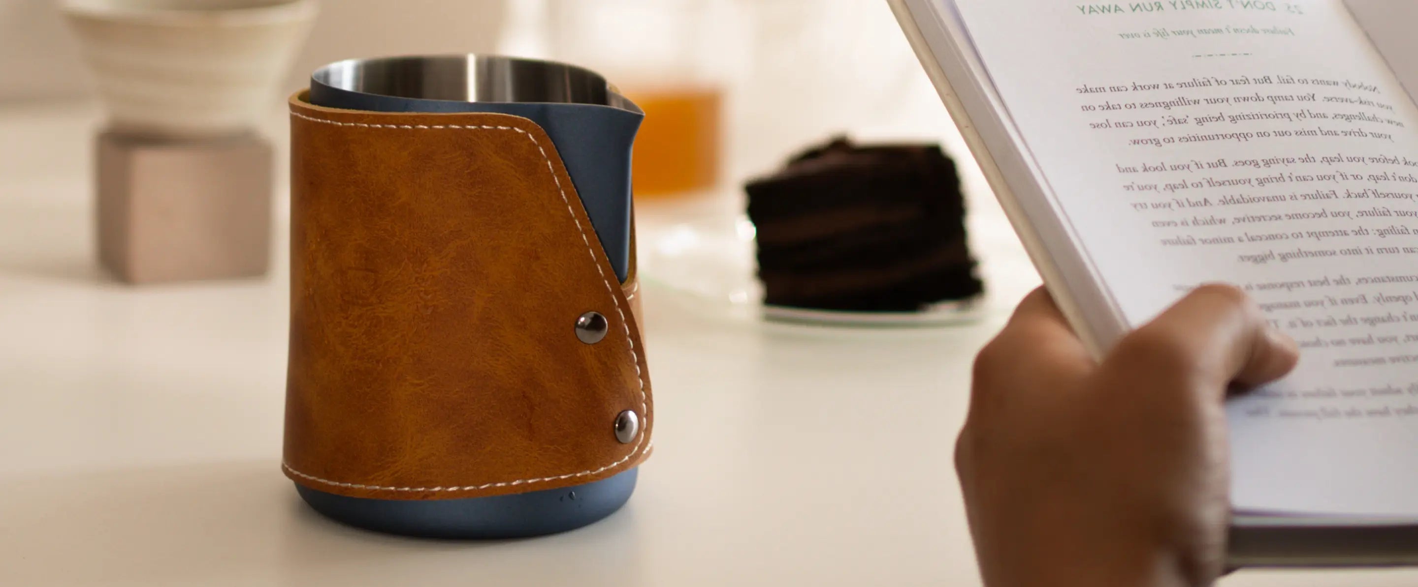 Brista Milk Pitcher placed on a table with a book, featuring a sleek tan leather grip and durable design.