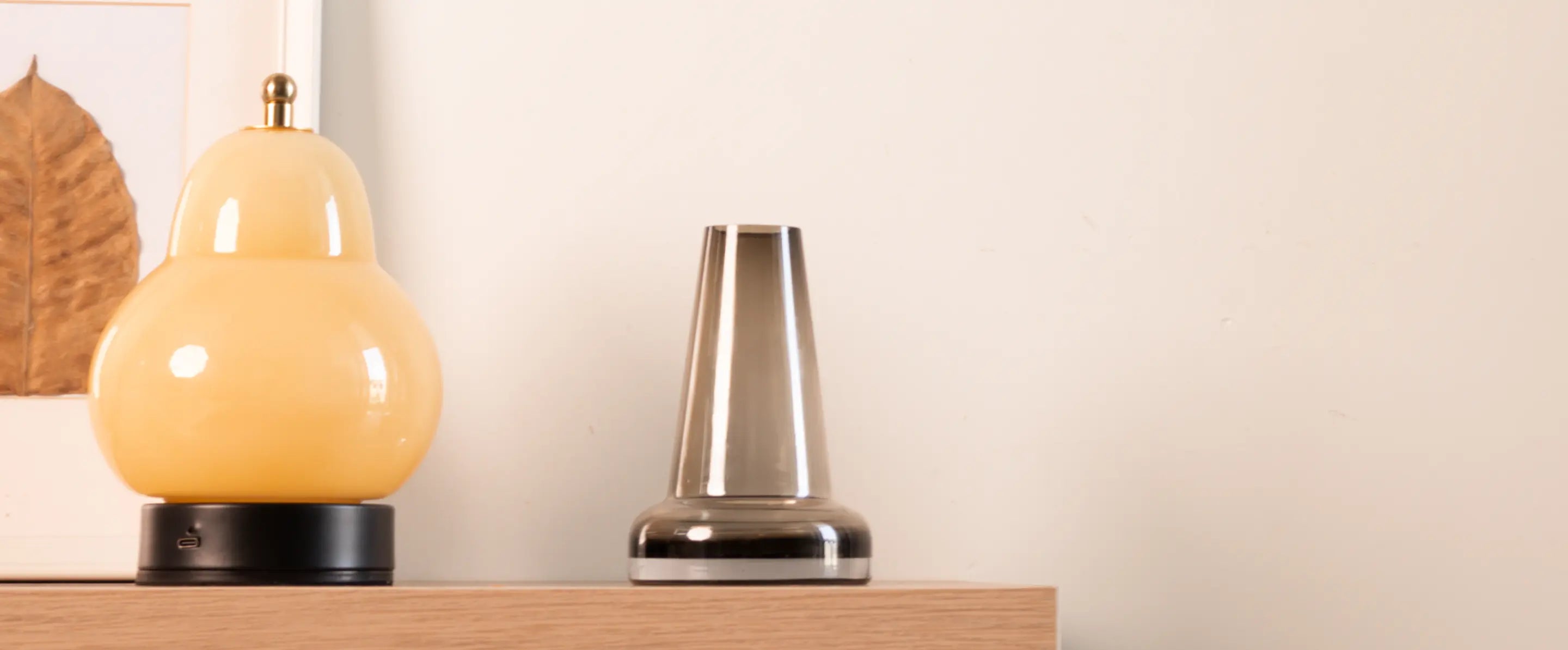 close-up of the Pitcher Glass Vase - Long Neck placed on a wooden shelf, next to a decorative yellow lamp and framed art