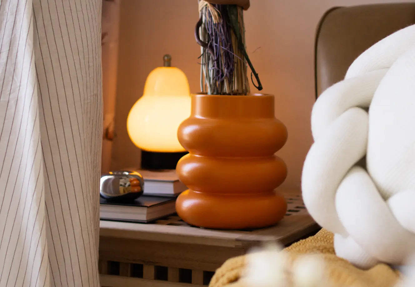 Modern home decor setup featuring a ceramic orange vase, cozy white cushion, ambient lamp, and stacked books on a wooden console.