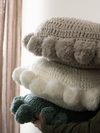 Stack of round, knitted pom-pom cushion in neutral tones — grey, white, and blue, styled in natural light for a cozy, modern look.