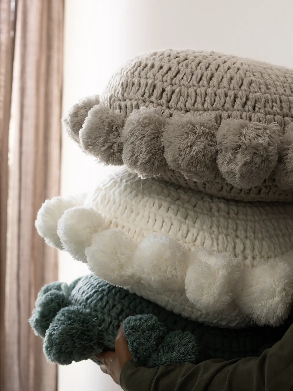 Stack of round, knitted pom-pom cushion in neutral tones — grey, white, and blue, styled in natural light for a cozy, modern look.