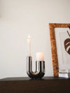 Modern chrome U-shaped candle holder with taper and pillar candles styled on a dark wooden console shelf.