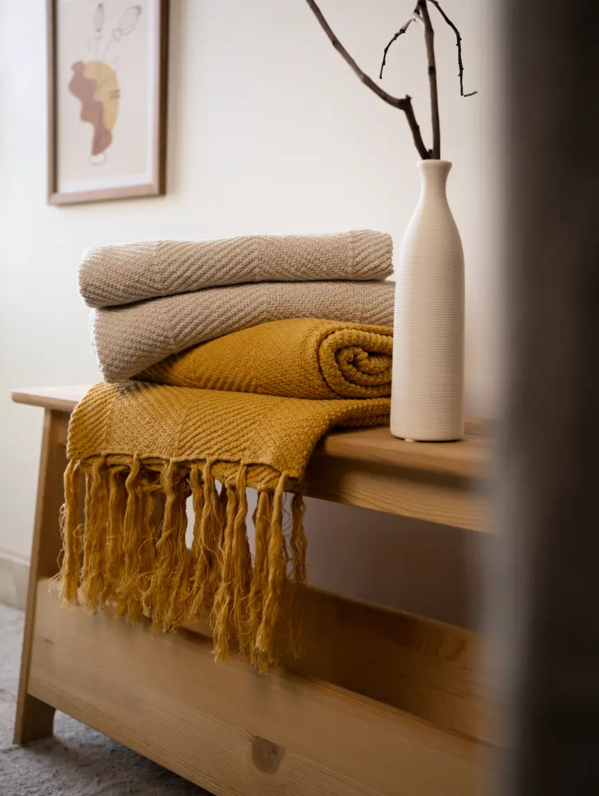 Stack of knitted throw blankets in yellow and beige on a wooden surface with a blurred background.