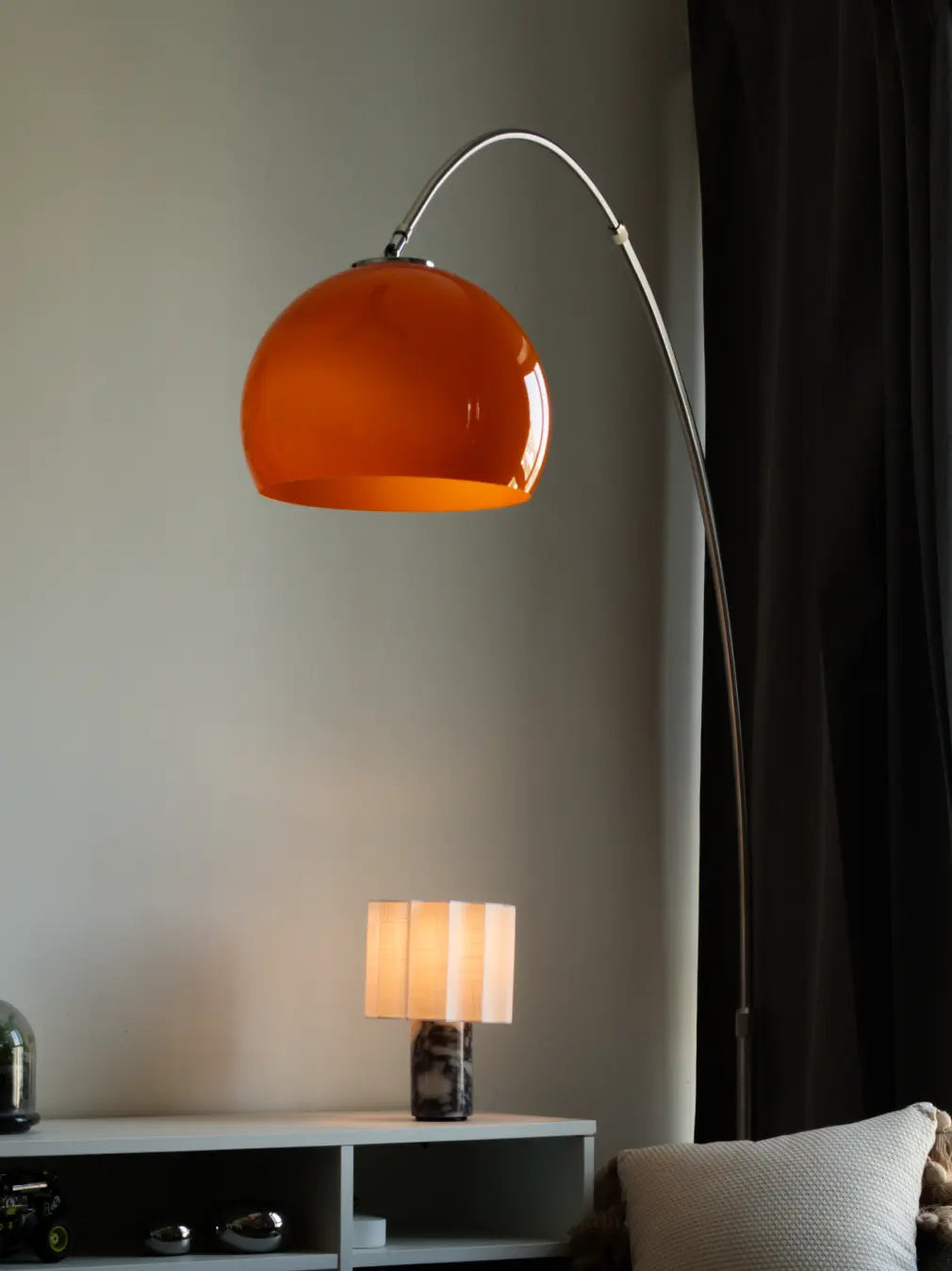 An orange glass dome floor lamp with a curved silver metal stand, shown in a modern living room setting next to a black curtain.