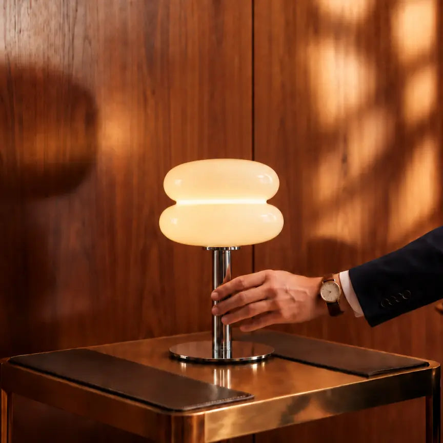 A modern two-tiered table lamp with a glowing warm white glass shade and a polished silver metal stem, shown on a metallic side table with a person's hand reaching toward it against a warm wood-paneled background.