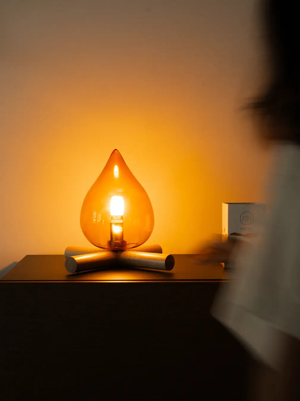 Campfire Glass table lamp shaped like a teardrop on a wooden surface with a blurred person in the foreground.