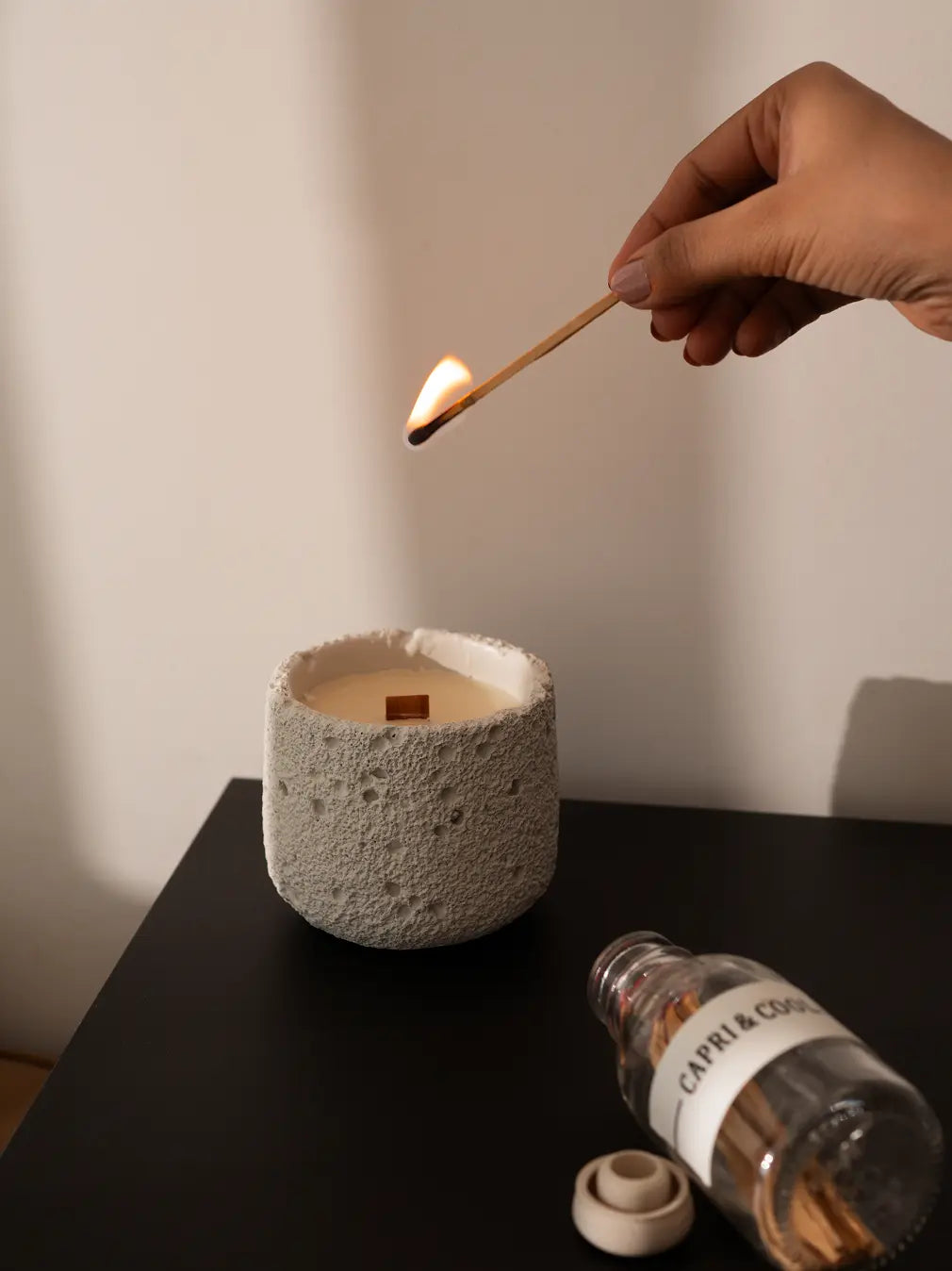 Lighting white Capri Candle with a matchstick, featuring raw concrete jar and wooden wick