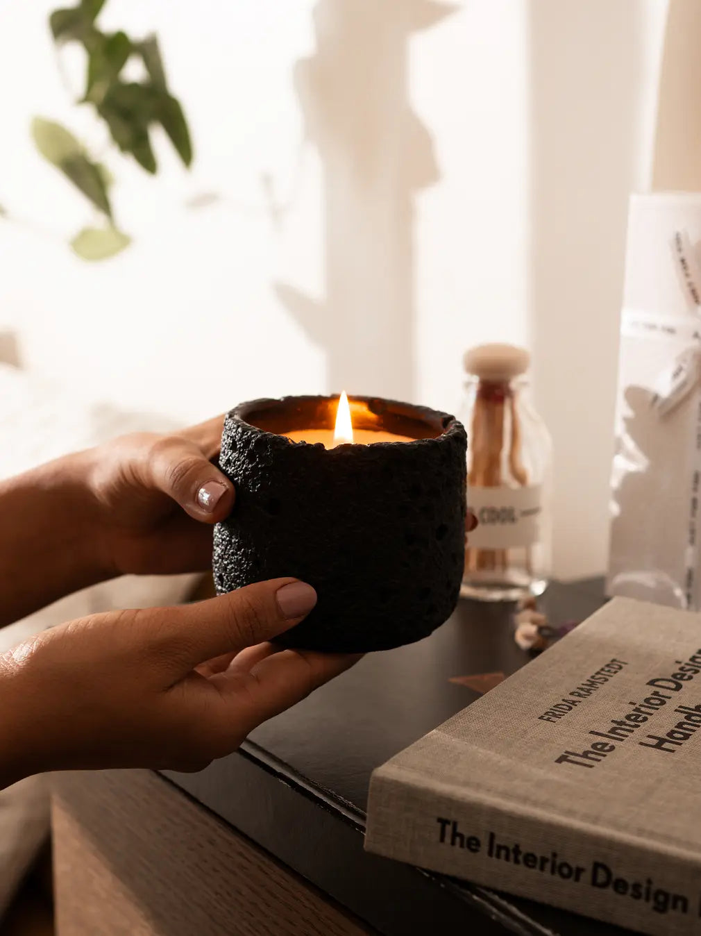 Hands holding black Capri Candle with soft flame, placed beside home decor accessories