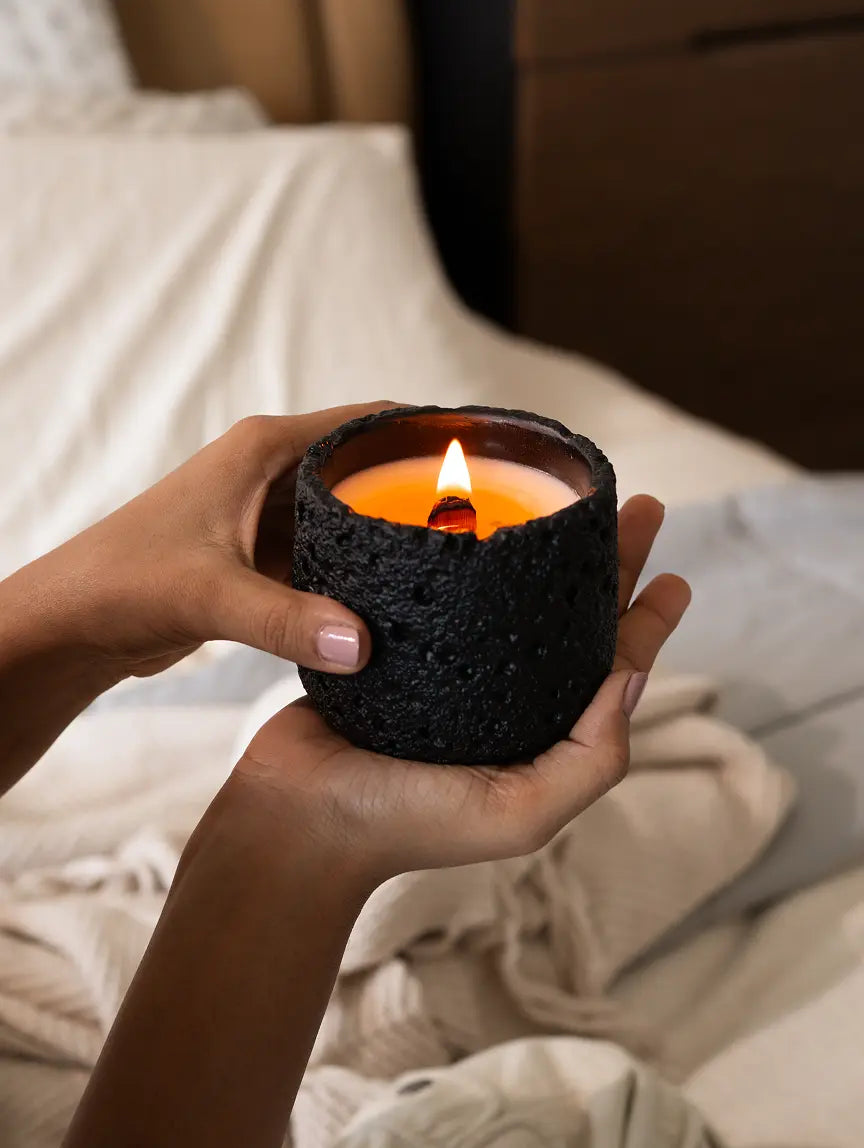 Hands holding black Capri Concrete Scented Candle on bed, soy wax with wooden wick