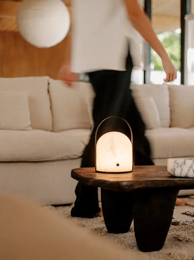 A glowing marble-look dome lamp with a black metal frame sits on a dark wooden coffee table in a modern living room. A person is blurred in the background walking past a cream-colored sofa.