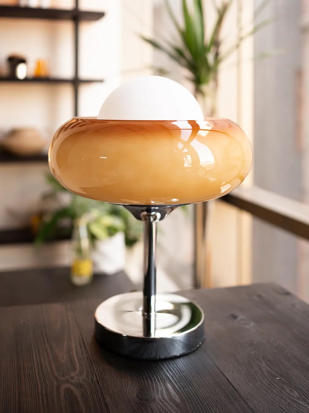 Dawn Glass Table Lamp with orange-brown glass shade and silver base glowing softly on a modern wooden table near a window.