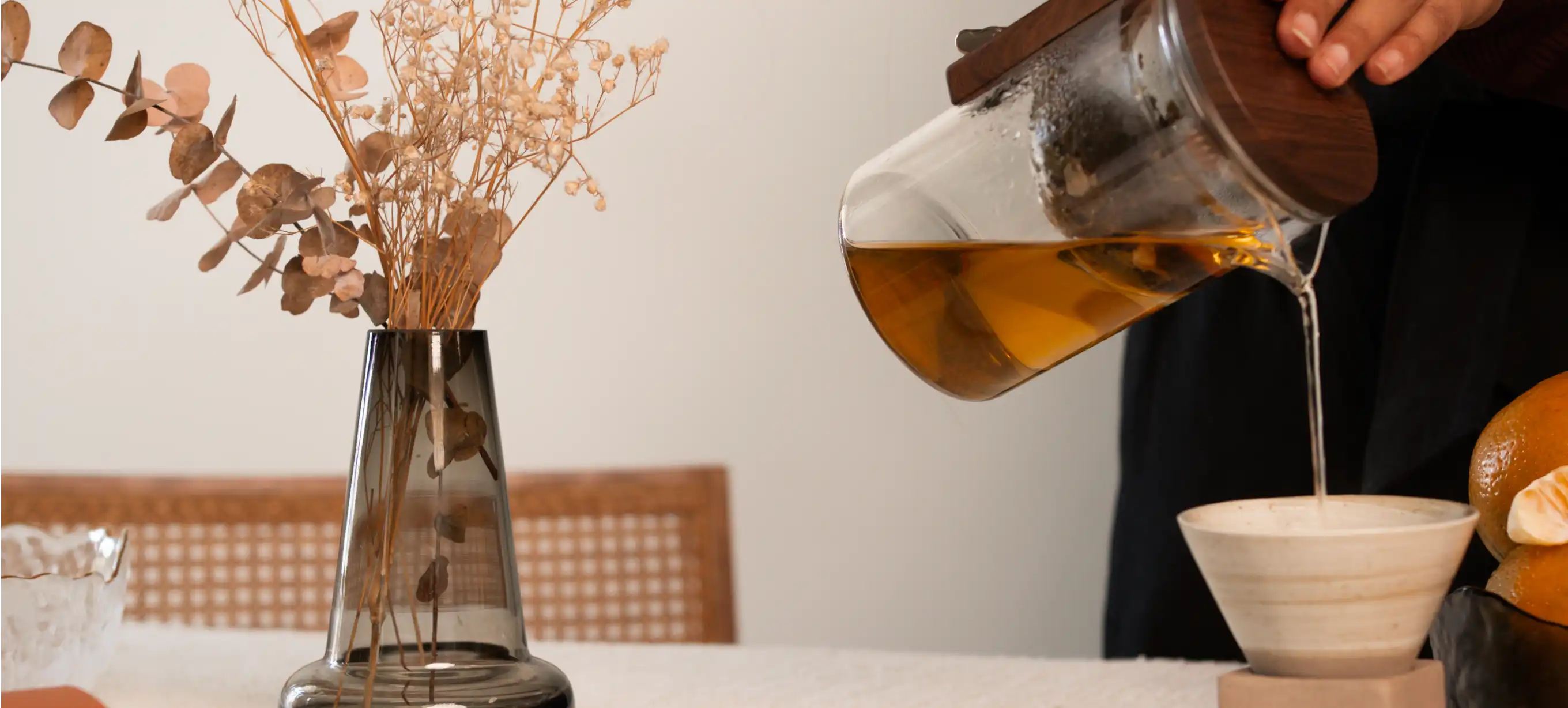 Person pouring tea from a glass teapot into a white ceramic cup with a blurred background