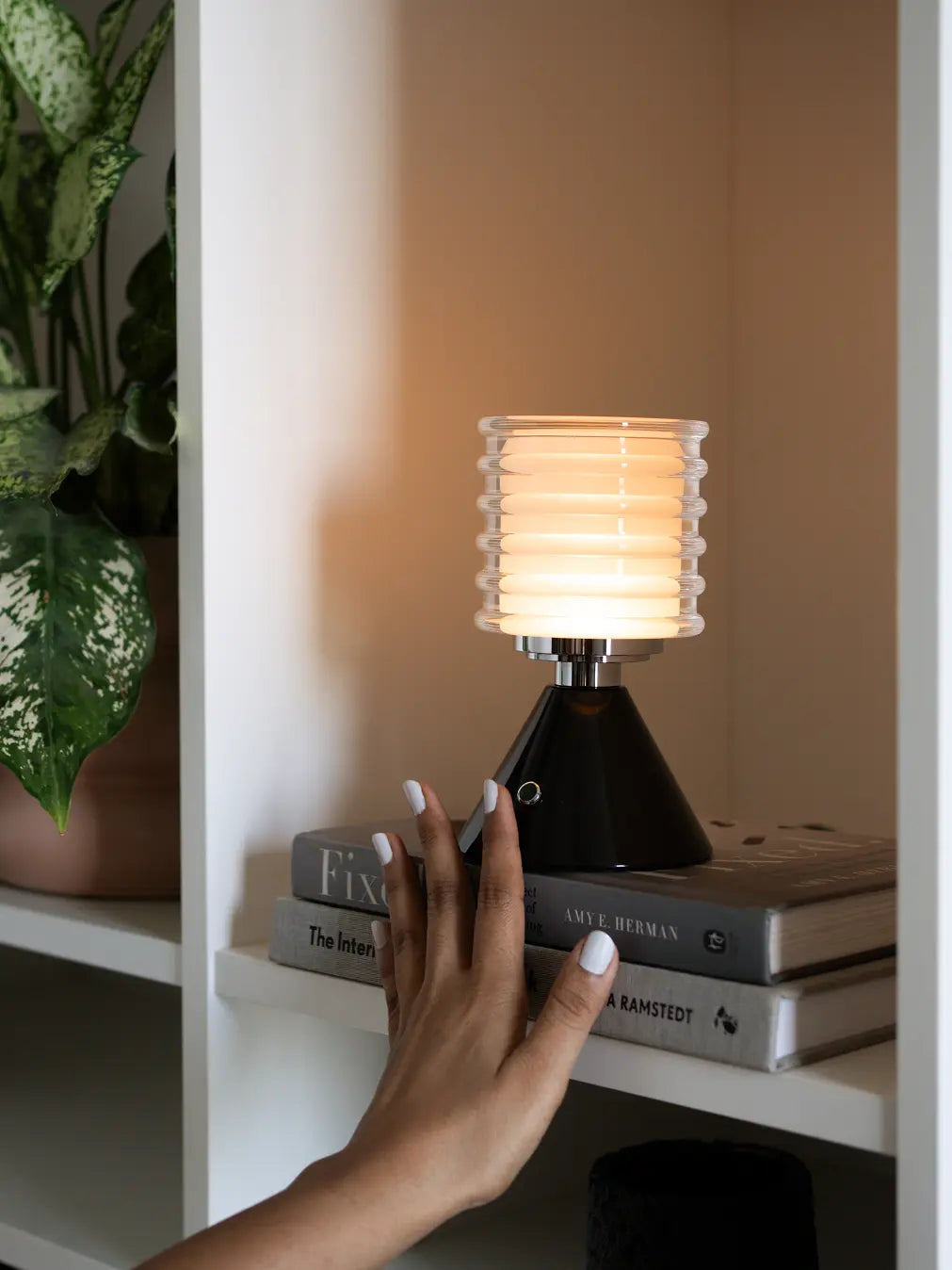 Dusk table lamp on a stack of books with a hand reaching towards it, in a cozy indoor setting.
