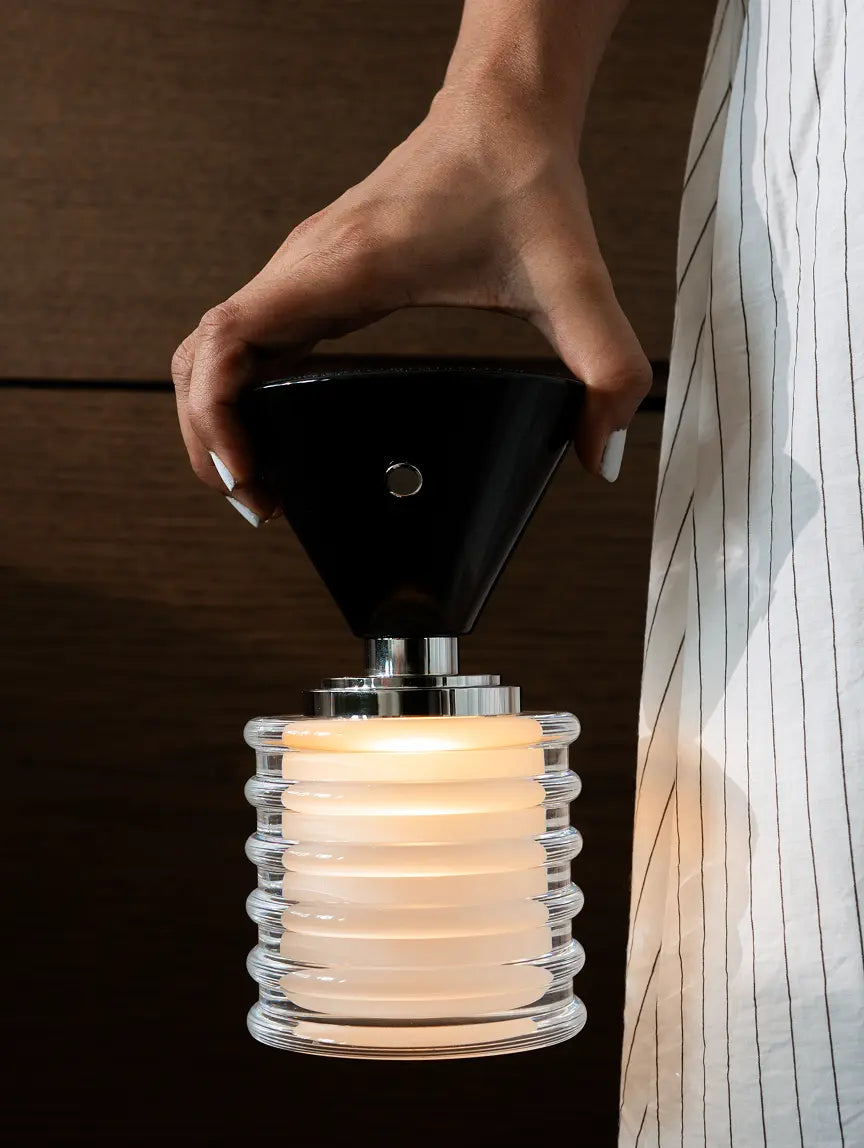 Hand holding a black lampshade over a clear glass lamp with a warm glow.