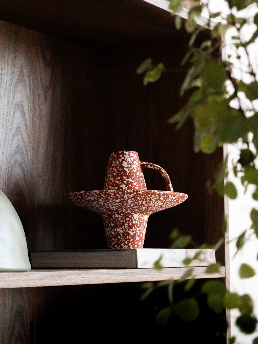 Speckled ceramic flower vase on a wooden shelf with greenery in the foreground