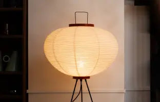 Lamp with a round paper shade on a tripod base in a minimalistic setting.