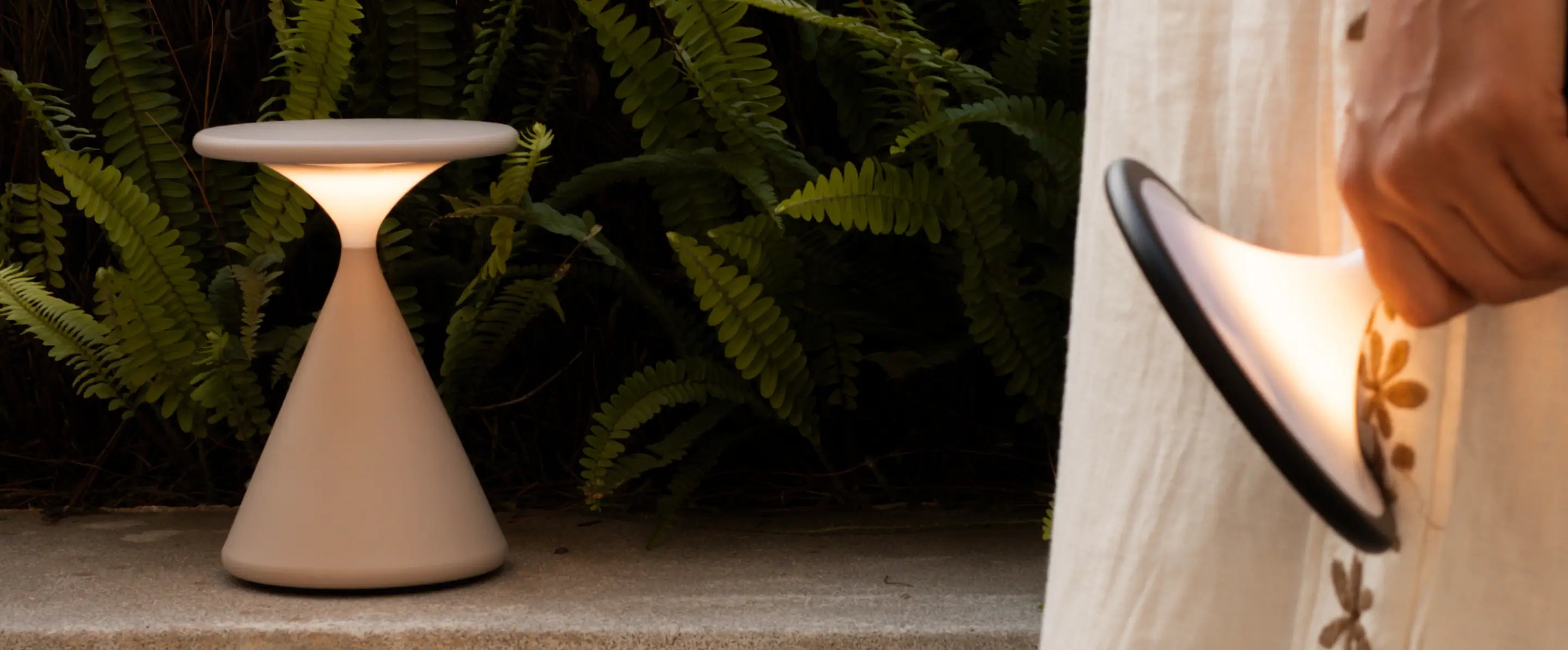 Flux Portable Table Lamp in bone and black variant shown in an outdoor greenery setup.