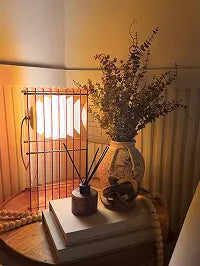 Cozy home decor with a glowing table lamp, dried foliage in a rustic vase, reed diffuser and stacked books on a wooden side table.