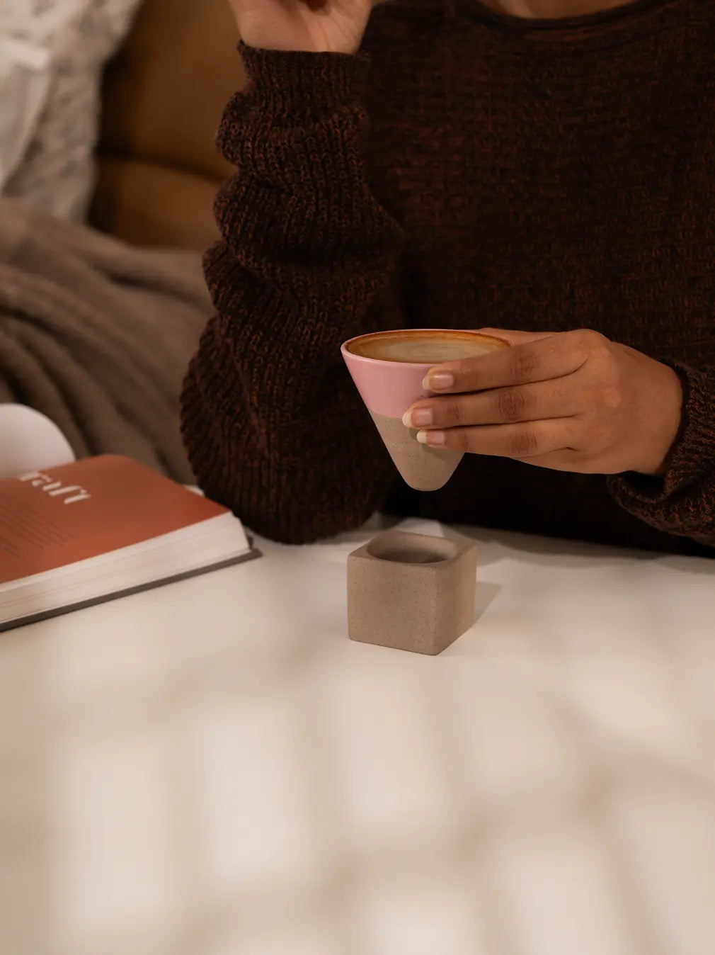 Person holding a pink ceramic cup with a book kept aside