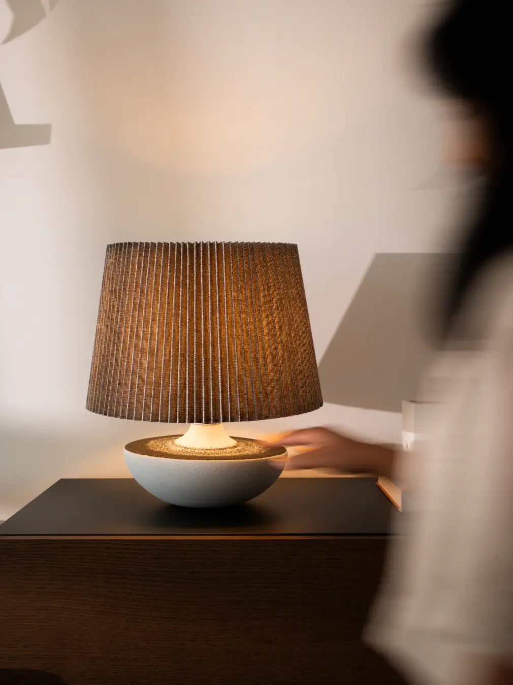 Wabi Sabi Table lamp with pleated brown shade on a dark surface, blurred person in the background