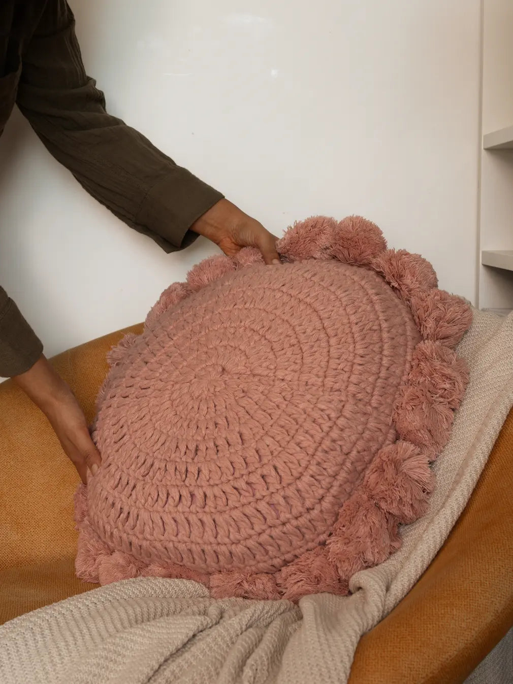 Person holding a pink crochet cushion on a chair.