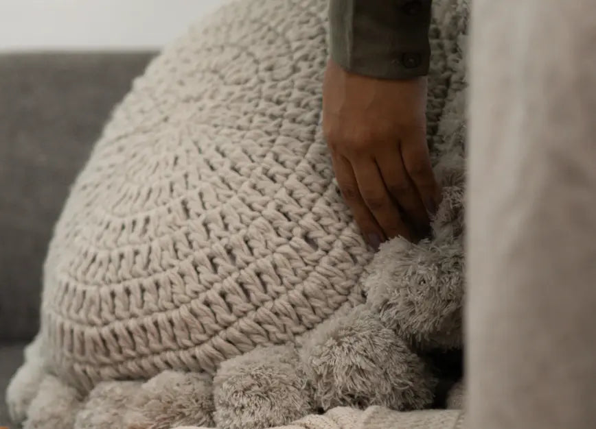 Hand adjusting Gharko's fluffy round cushion with tassel on a couch.