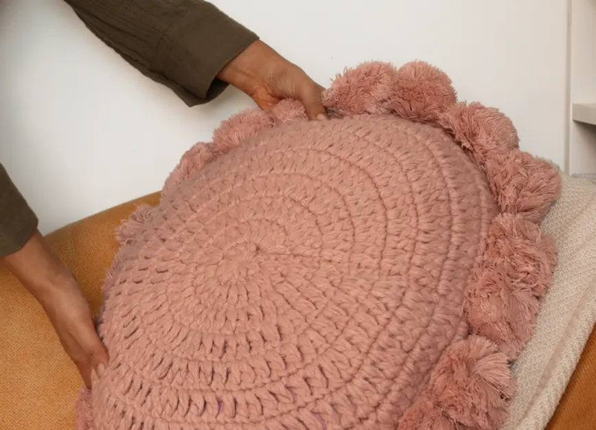 Pink textured cushion with pom-poms being held by a person on a chair
