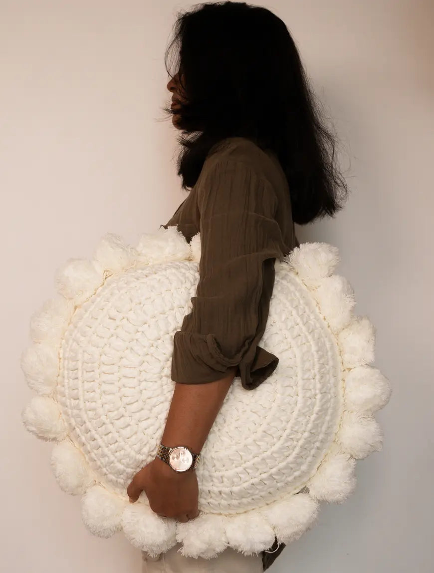 Person holding on a round white cushion with pom-poms against a plain background