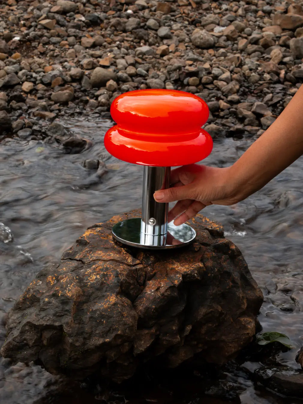 Orbit Red portable table lamp with chrome base placed on a rock beside a stream