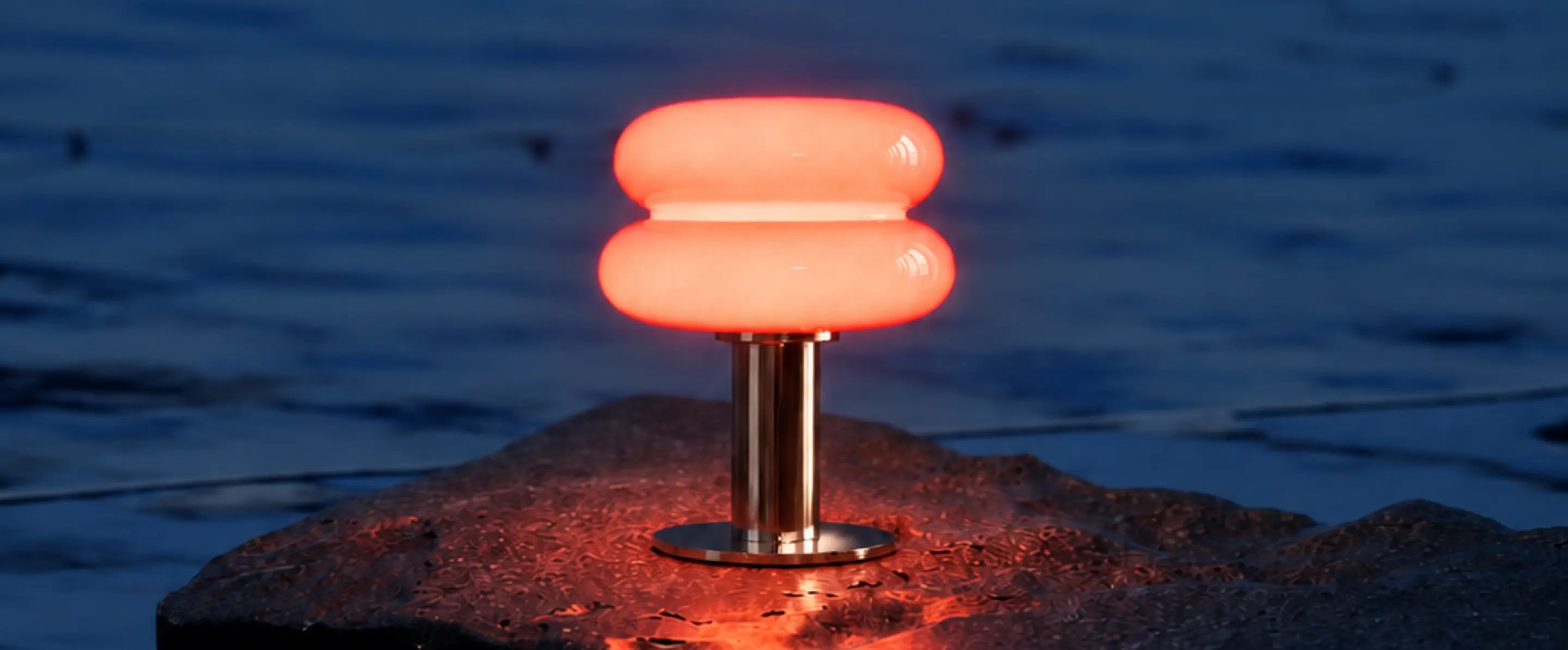 Orbit Red portable table lamp glowing on a seaside rock at dusk
