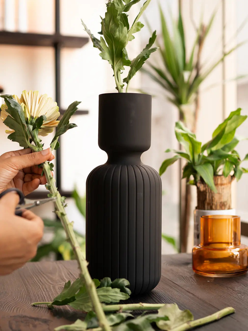 Person arranging flowers around a ribbed black vase on a wooden surface with plants in the background.