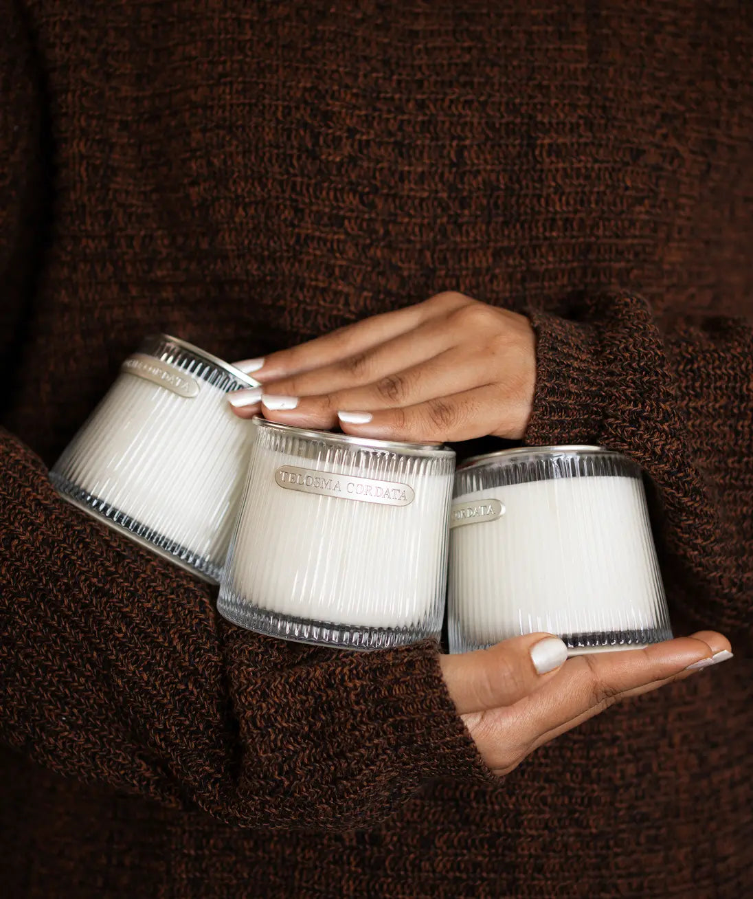 Three white scented candles in ribbed clear glass jars are held in a person's hands against a dark brown knit sweater background. The center jar has a visible label reading 'candle'.
