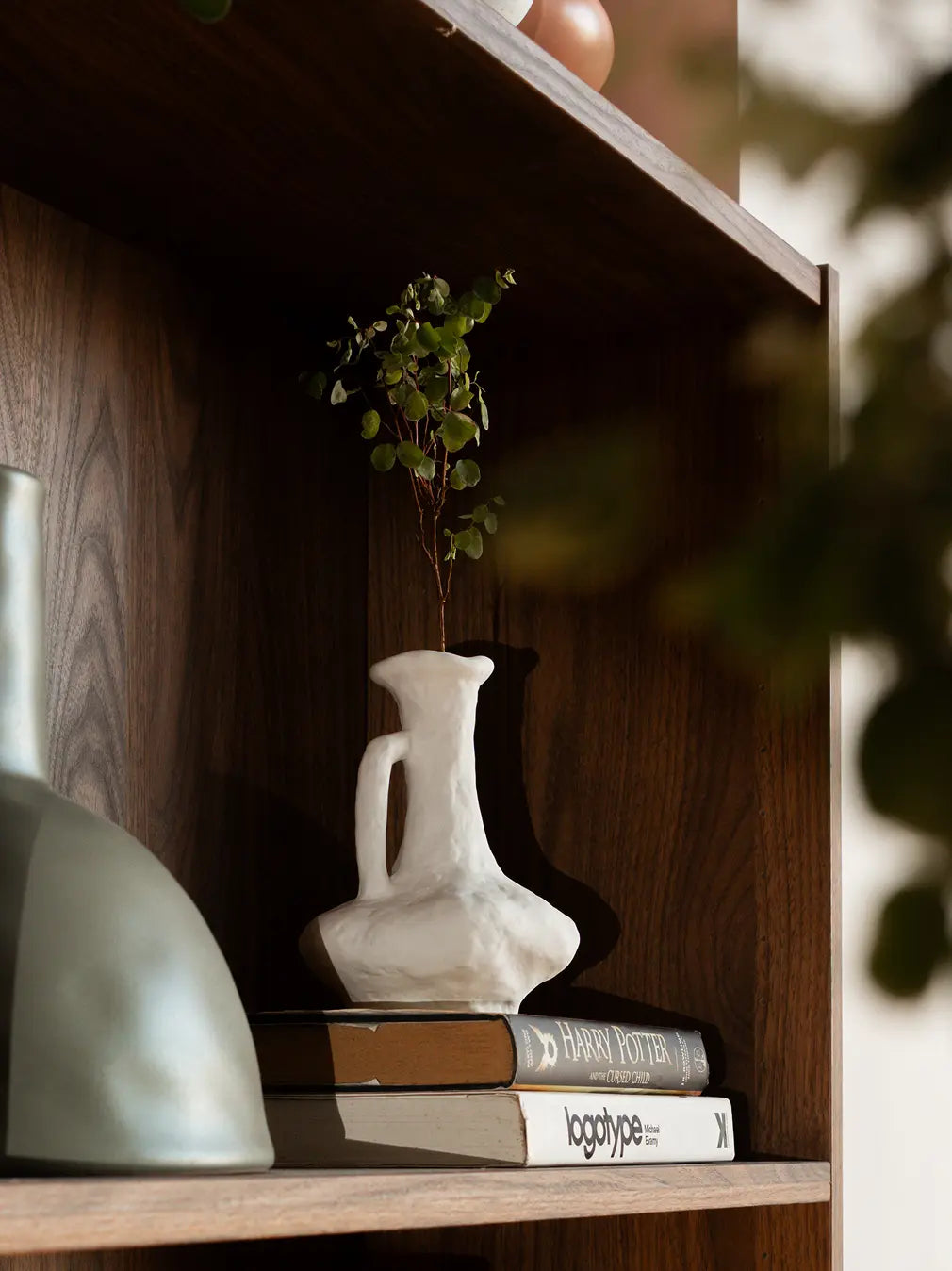 White ceramic flower vase with handle placed on wooden shelf, styled with dried stems in warm light.