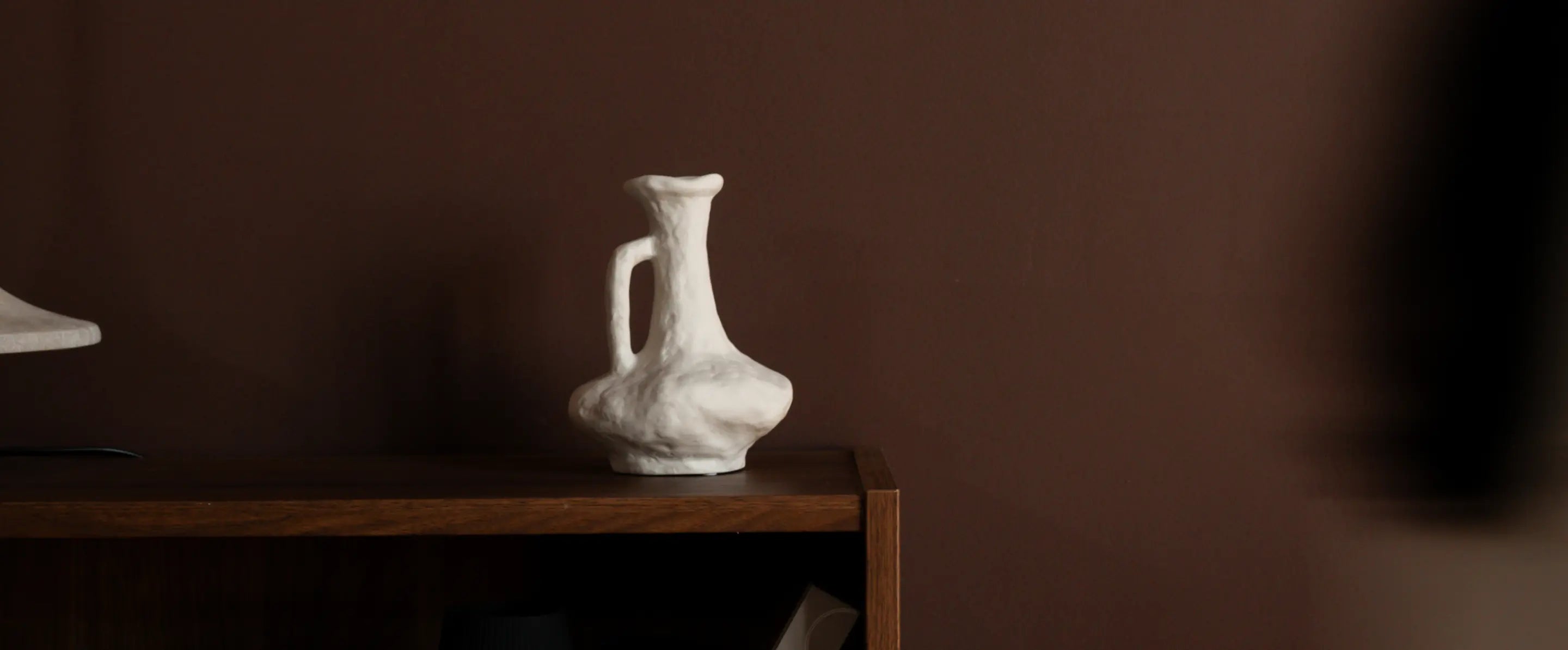 Suki Classic Ceramic Vase placed on wooden shelf against warm brown wall.