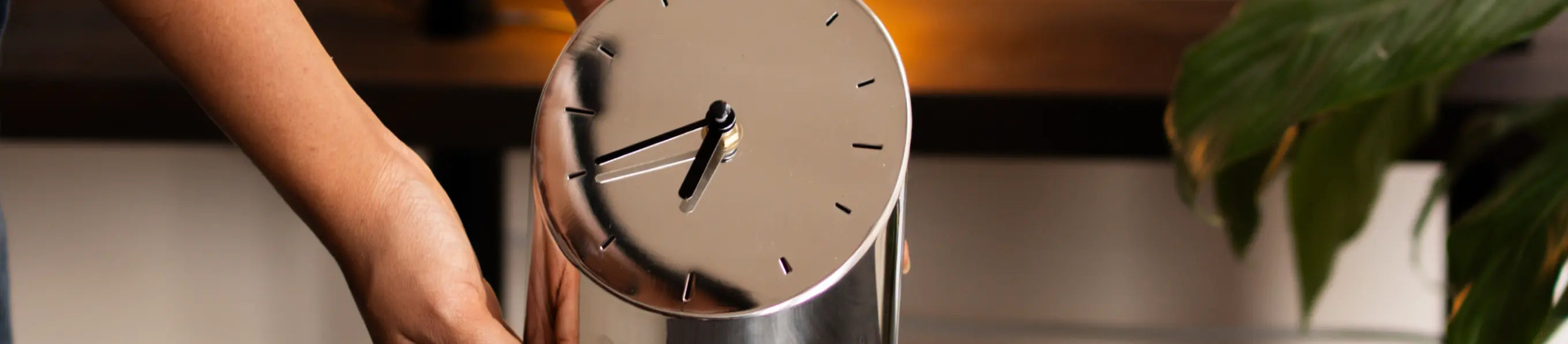 Person placing a modern silver metal table clock on a wooden desk near warm lighting.