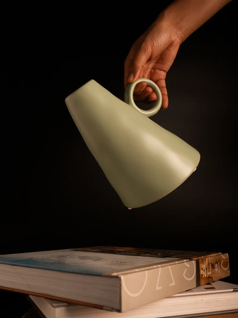 Hand holding a light green ceramic vase above a book on a dark background