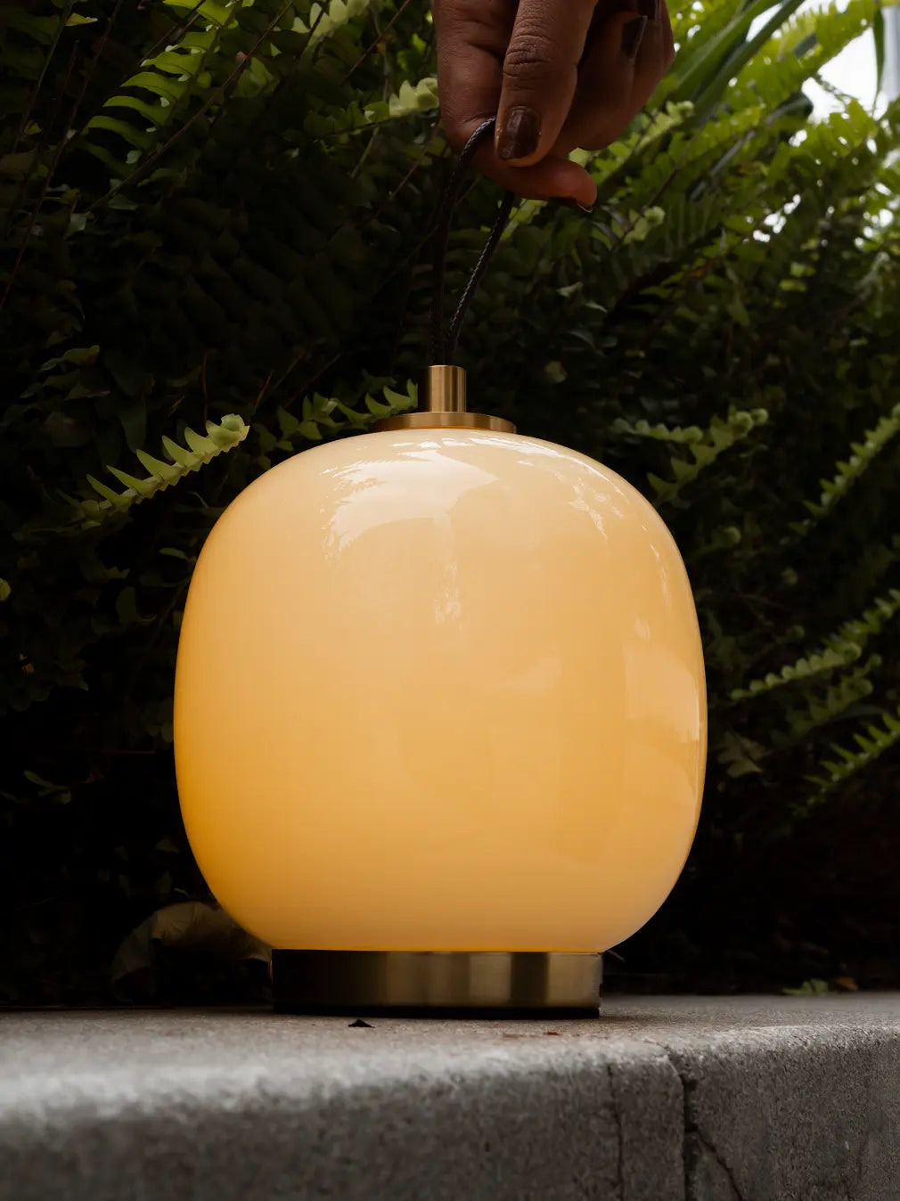Light yellow Dew portable LED lamp glowing outdoors with leather carry strap